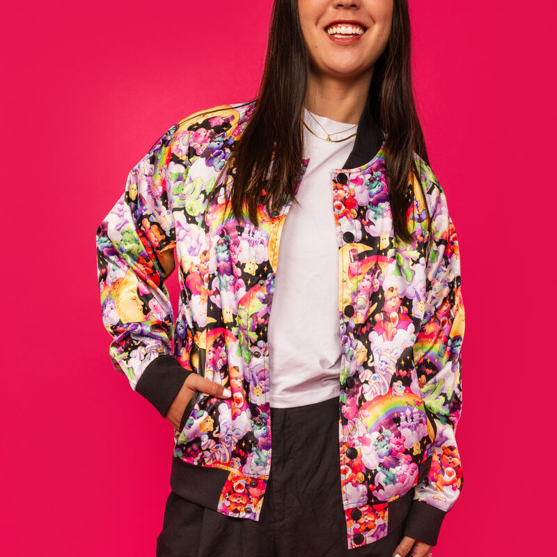 Woman standing against a bright pink background wearing the satin Loungefly Care Bears x Universal Monsters bomber jacket.  