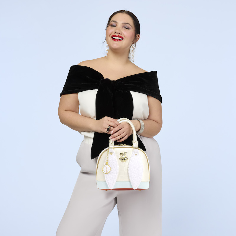Woman smiling at camera wearing white pants and a white-and-black off-the-shoulder top. She wears the Loungefly Alice in Wonderland White Rabbit Handbag over one arm. 
