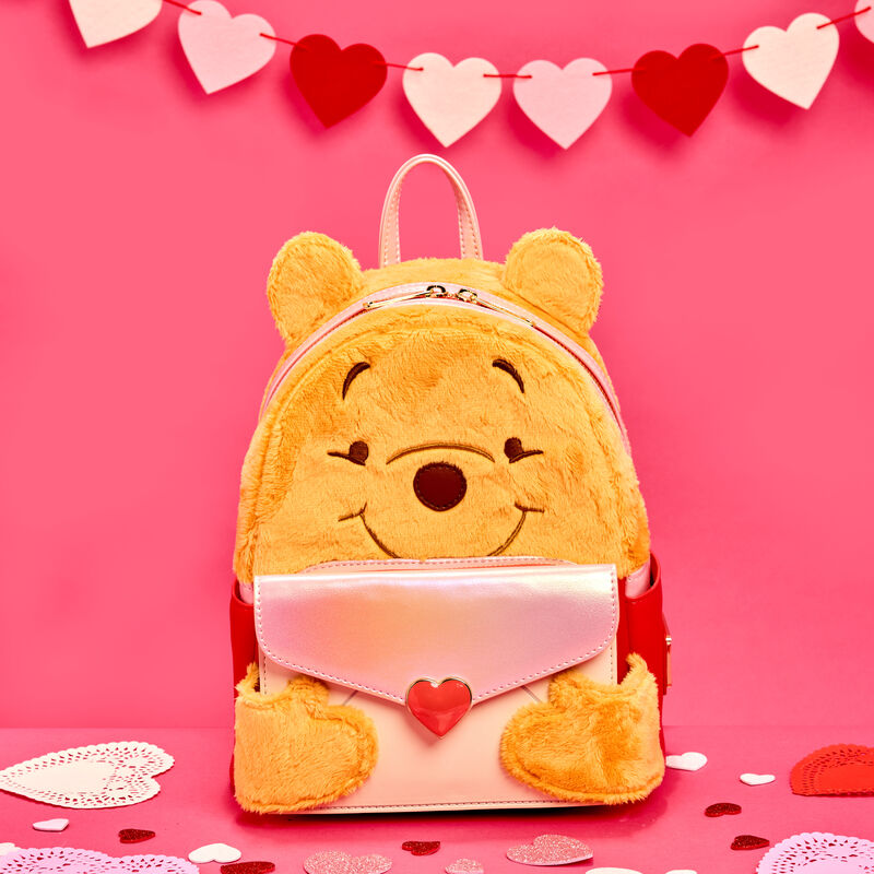 Plush Loungefly Winnie the Pooh mini backpack featuring Pooh holding an envelope with a heart on it. The bag sits against a pink background that has heart-shaped doilies spread around and a heart garland hung up. 