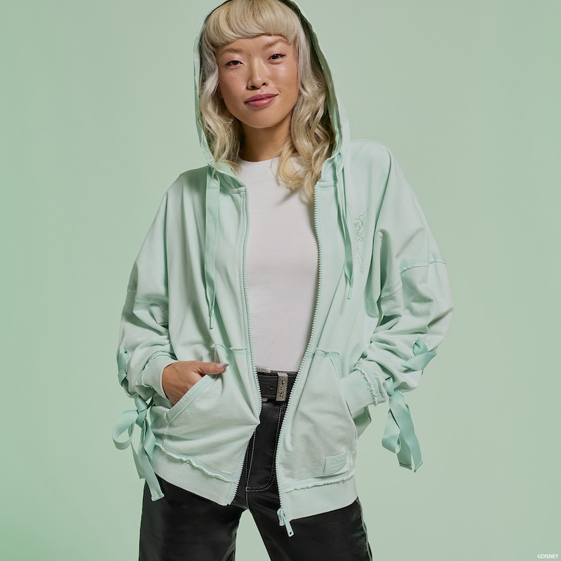 Woman smiling at camera wearing the light green Loungefly Peter Pan Tinker Bell Ribbon Zip Up Hoodie. The hoodie is unzipped over a white shirt and black jeans, and the woman poses with her hands in the hoodie pockets. She stands against a light green background. 