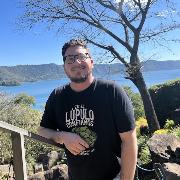 Man in glasses standing outside,  leaning against a wooden railing with a lake in the background