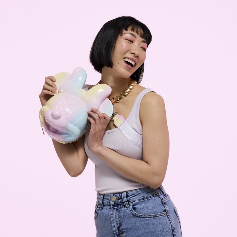 Woman smiling and holding up the Loungefly Minnie Mouse Pastel Tie-Dye Molded Figural Crossbody bag. She wears a tank top and jeans and stands against a pink background. 