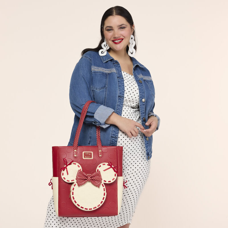 Woman facing camera and smiling, wearing a white dress with black polka dots and denim jacket. She wears the Loungefly Minnie Mouse Rock the Dots Tote Bag over an arm. 