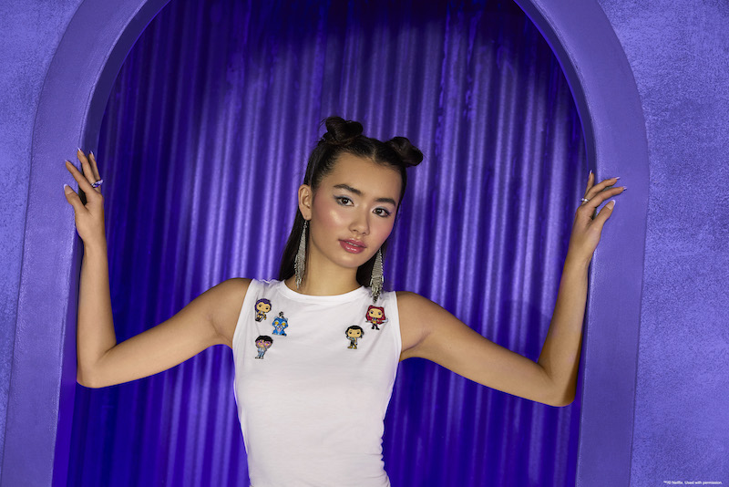 Woman standing against a purple background wearing a white, sleeveless top. She wears five of the six Loungefly KPop Demon Hunters Mystery Box Pins on her shirt: Rumi, Derpy, the Jinu chase, Mira, and Zoey. 