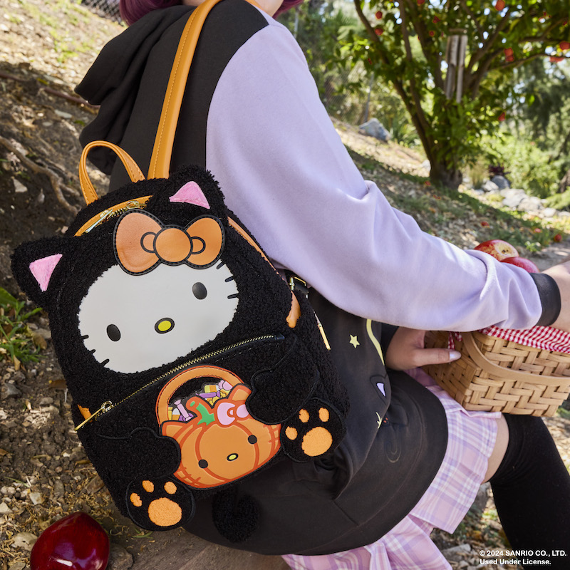 woman sitting outside wearing the Loungefly Sanrio Hello Kitty & Friends Costume Hoodie and wearing the plush Hello Kitty Costume Mini Backpack, which features Hello Kitty dressed as a black cat for Halloween.