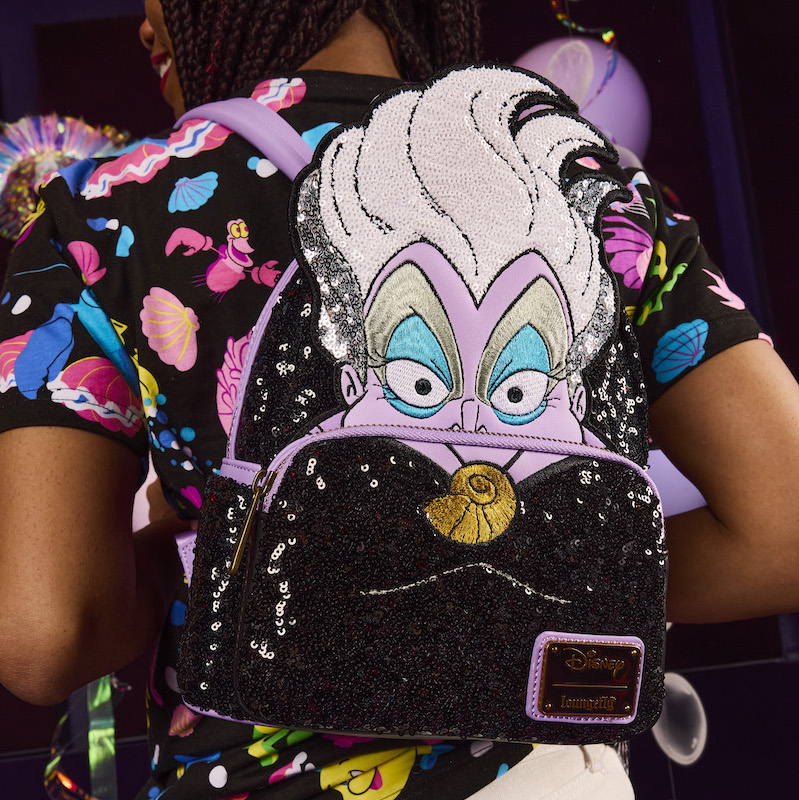 Woman facing away from camera wearing a black shirt with neon Little Mermaid characters on it, wearing the sequin Loungefly Ursula mini backpack, featuring Ursula in appliqué and sequin detail. 